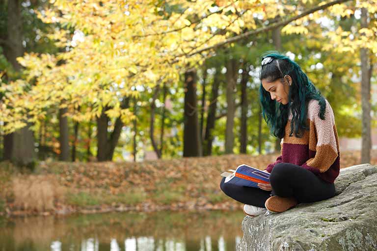 A student with brightly colored hair reads a book beside a stream.