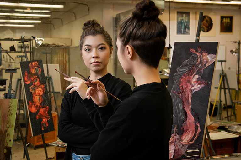 A student examines herself in the mirror as she prepares to paint a self portrait.